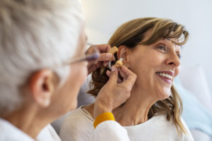 Doctor helping senior patient with hearing aid , close-up. Otolaryngologist putting hearing aid in senior woman's ear on light background. Adjusting of a hearing aid for an aged woman